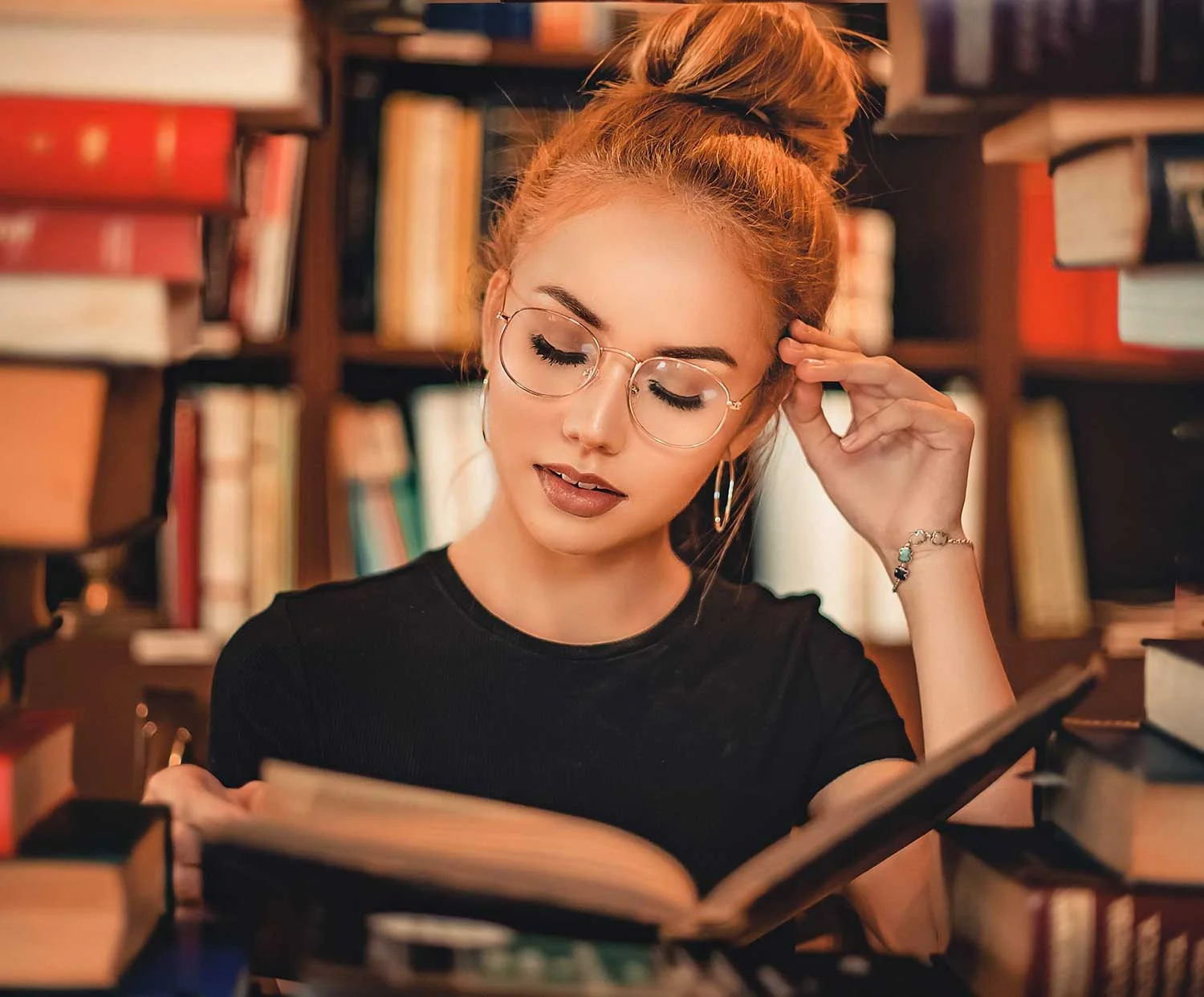 Rödhårig kvinna med glasögon, som läser en bok inne i ett bibliotek.